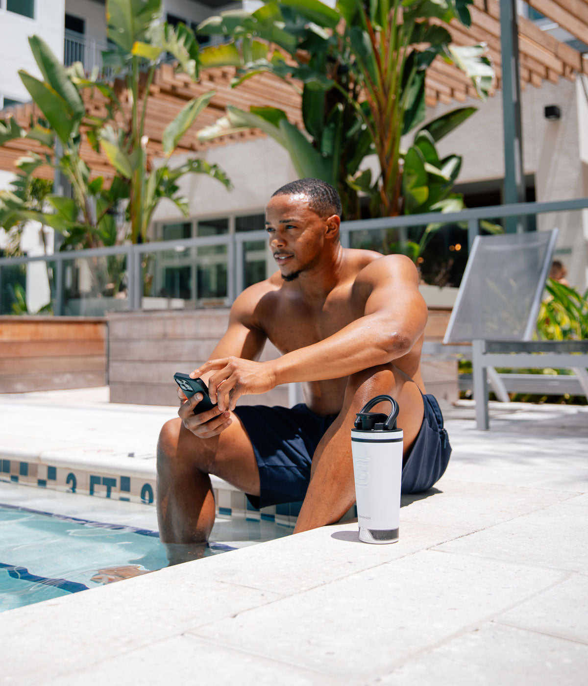 A young, African-American man is sitting by a pool with his feet in the water. He has his phone in his hand and a white 20oz Speaker Bottle next to him.