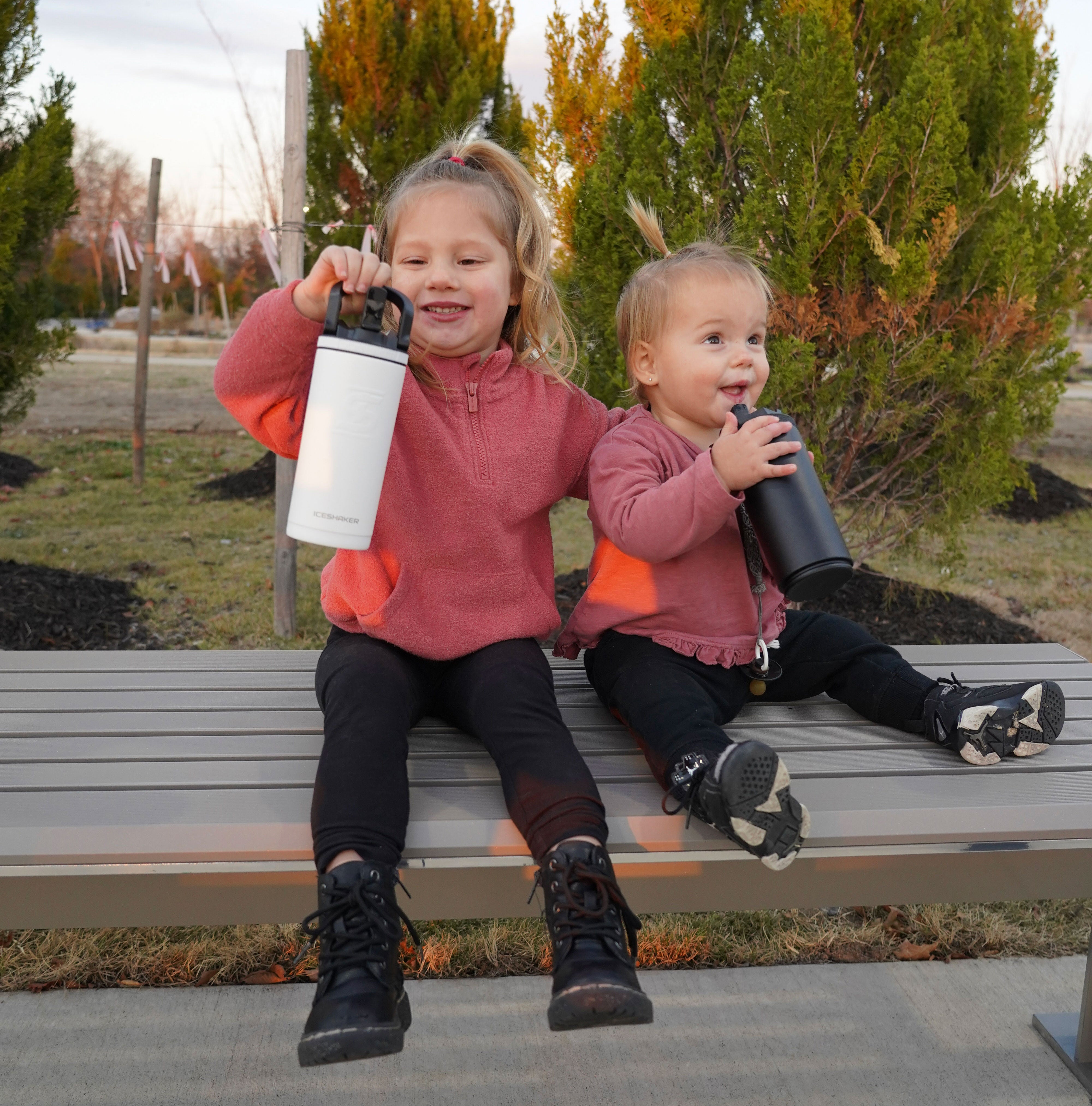 An image of two little girls sitting on a bench at a park. Both little girls are holding a 14oz Sport Bottle. One little girl is holding the bottle by it's handle and the other little girl is getting ready to take a drink from the Flip Up Sport Straw
