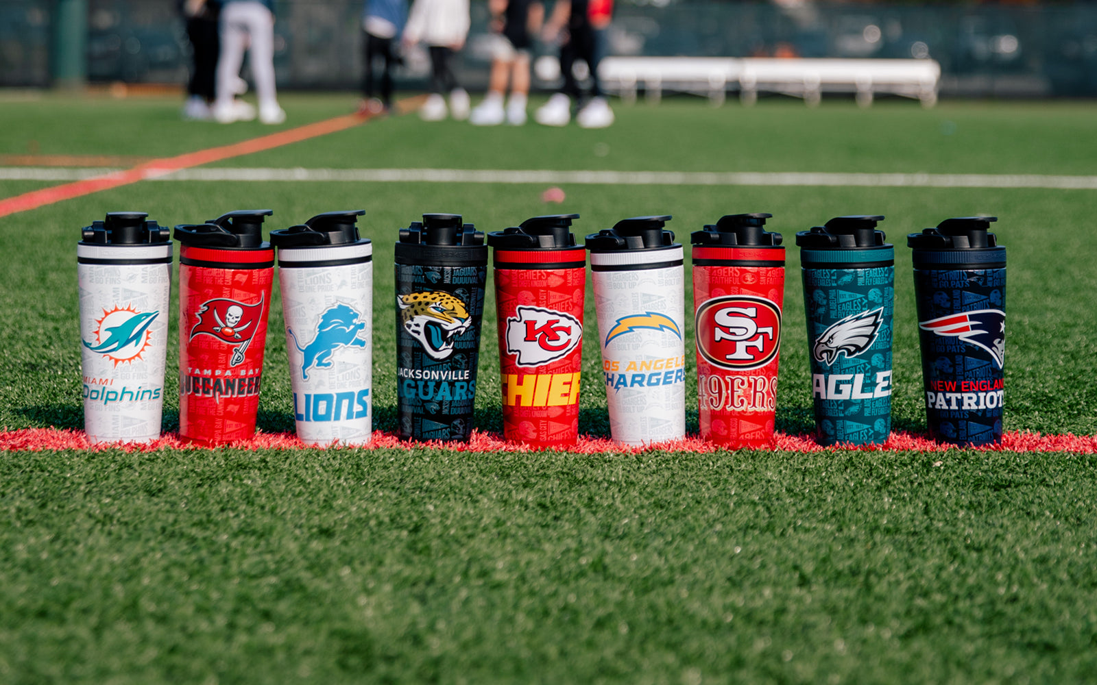 Various NFL 4D Ice Shakers lined up on a football field. People in the background are playing football.