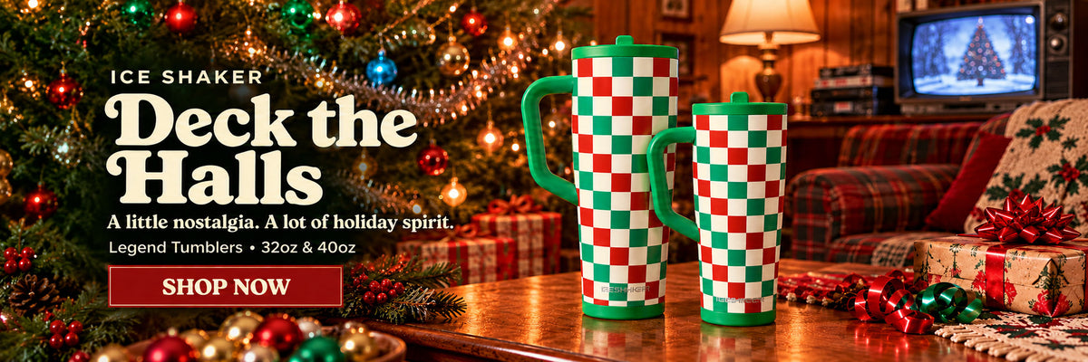 Two holiday-themed tumblers with a red and green checkered pattern and green handles are displayed on a wooden table in a cozy living room setting next to a decorated Christmas tree. Promotional text on the left reads 'ICE SHAKER Deck the Halls' and includes a 'SHOP NOW' button.