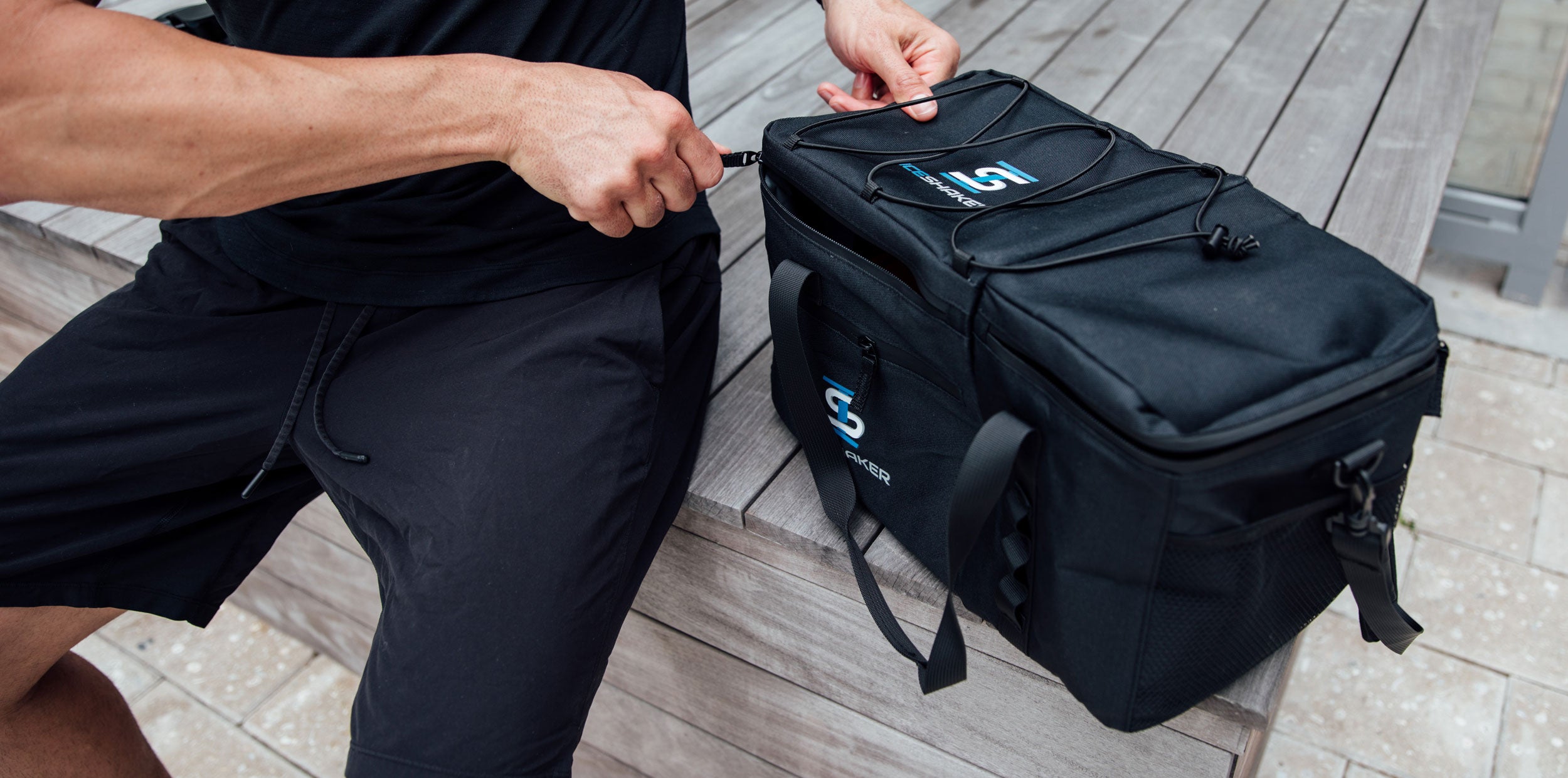 A close up image of the Ice Shaker's The Duffel Bag on a wooden platform. A man is sitting next to The Duffel and is opening the top of The Duffel with the zipper.