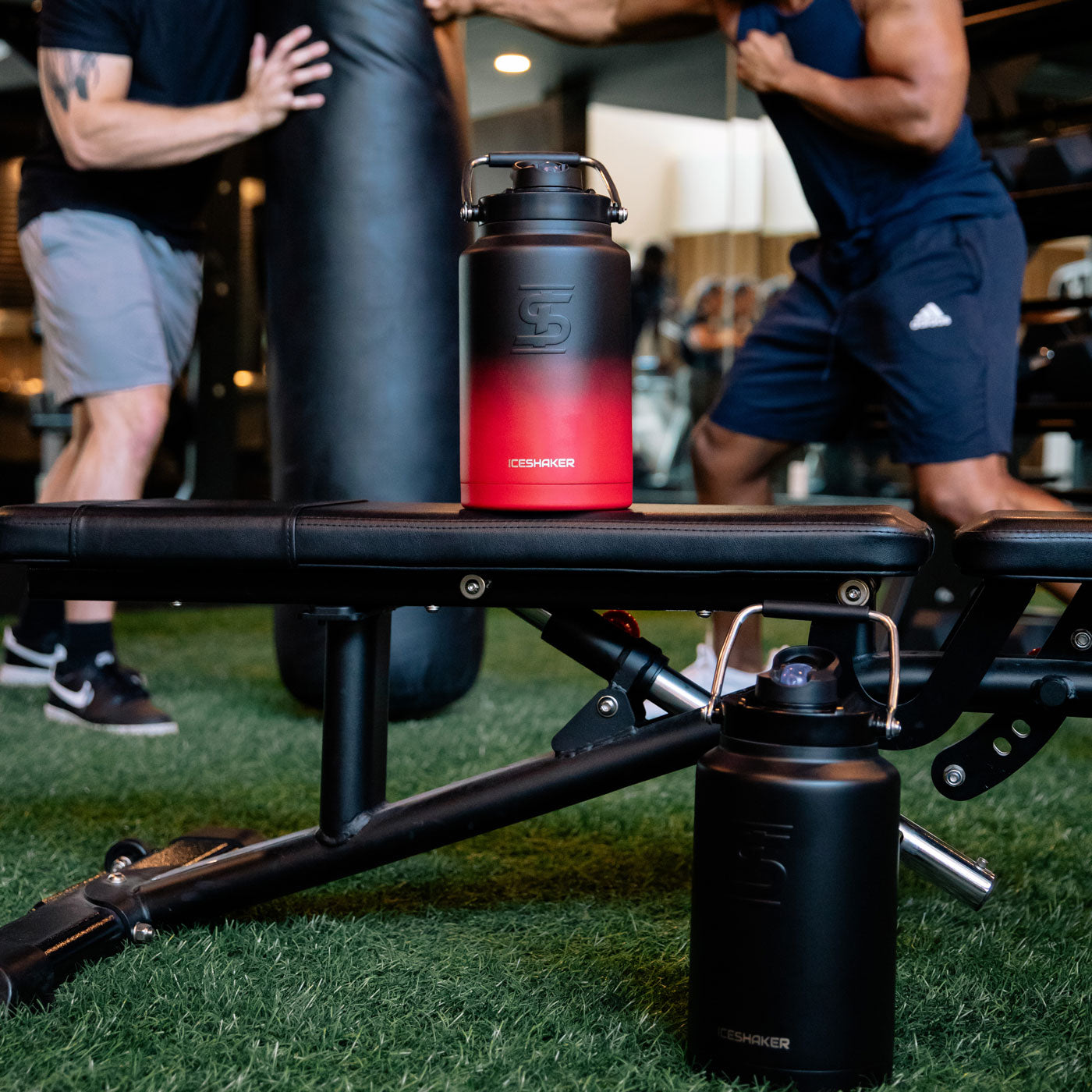 a black-colored One Gallon Jug and a Red Black Ombre-colored One Gallon Jug sitting on a bench in a gym. There are two men working out in the background.