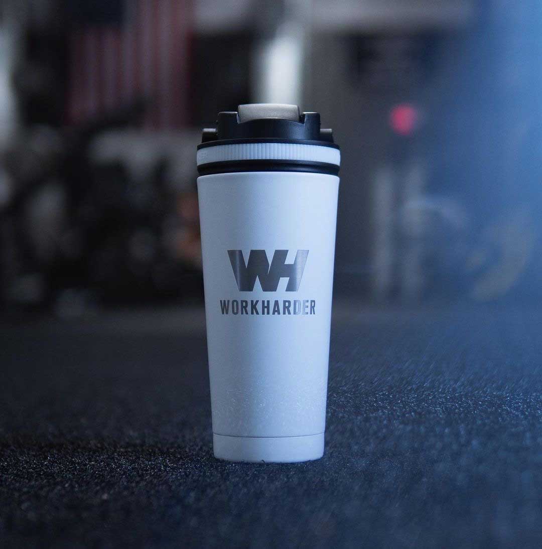 An image of the White 26oz Ice Shaker that's been customized with a Work Harder engraving. The Ice Shaker is sitting in a dark gym setting with the American flag in the background. Click now to shop All Engraved Bottles