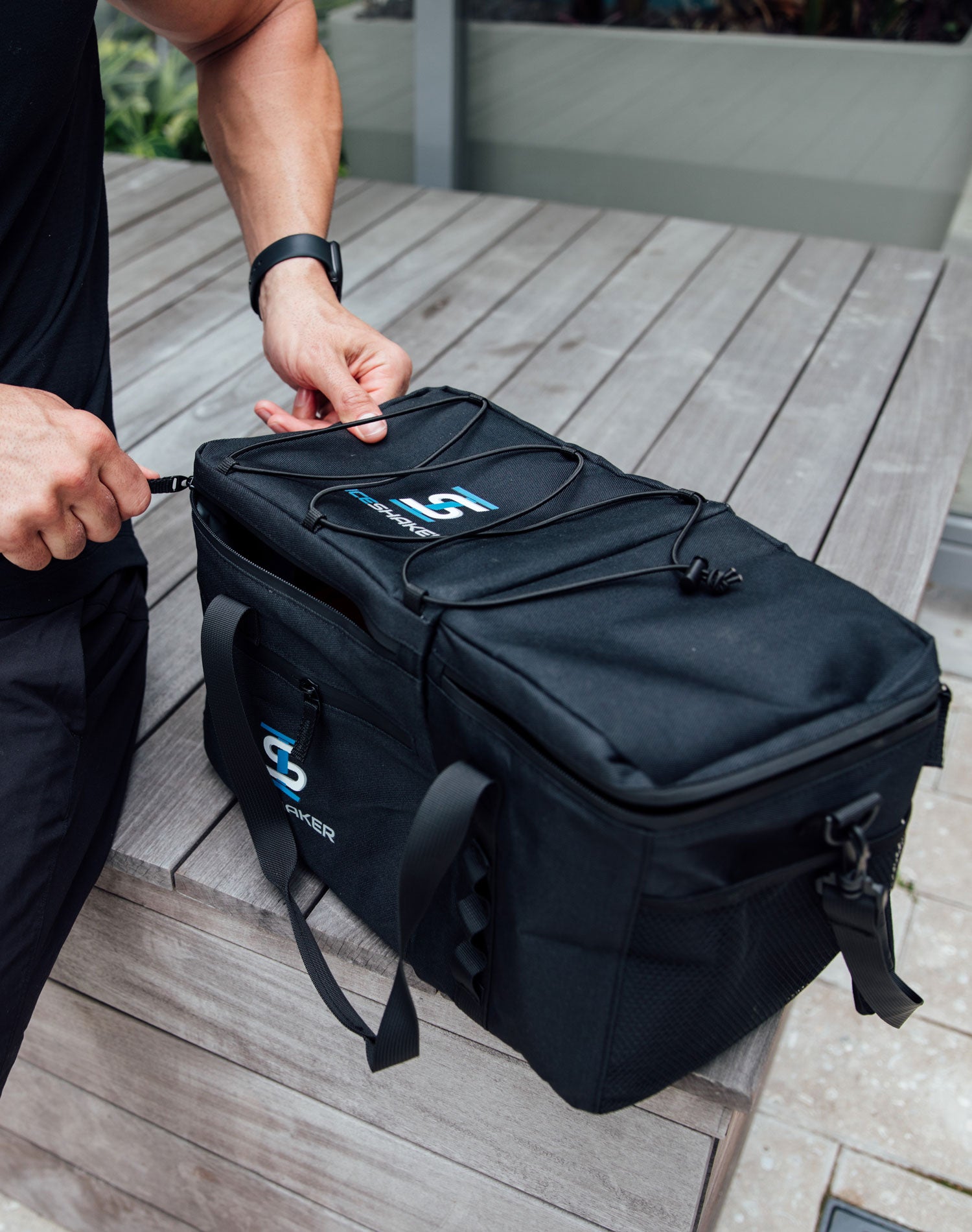 A close up image of the Ice Shaker's The Duffel Bag on a wooden platform. A man is sitting next to The Duffel and is opening the top of The Duffel with the zipper.