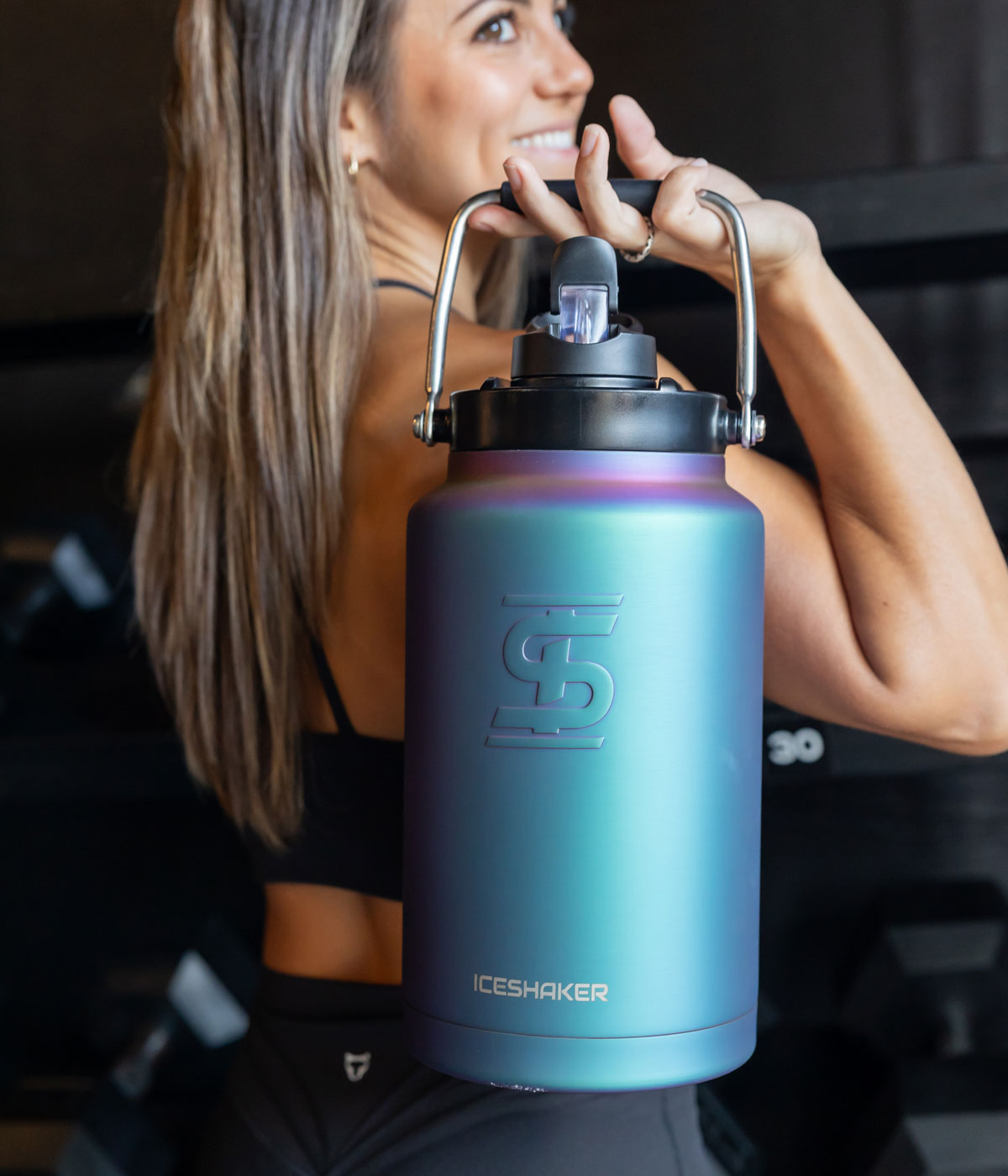 A woman with long brown hair, dressed in athletic clothing holds a wisteria-colored One Gallon Jug over her shoulder. She is holding the One Gallon Jug by its durable easy-carry handle.