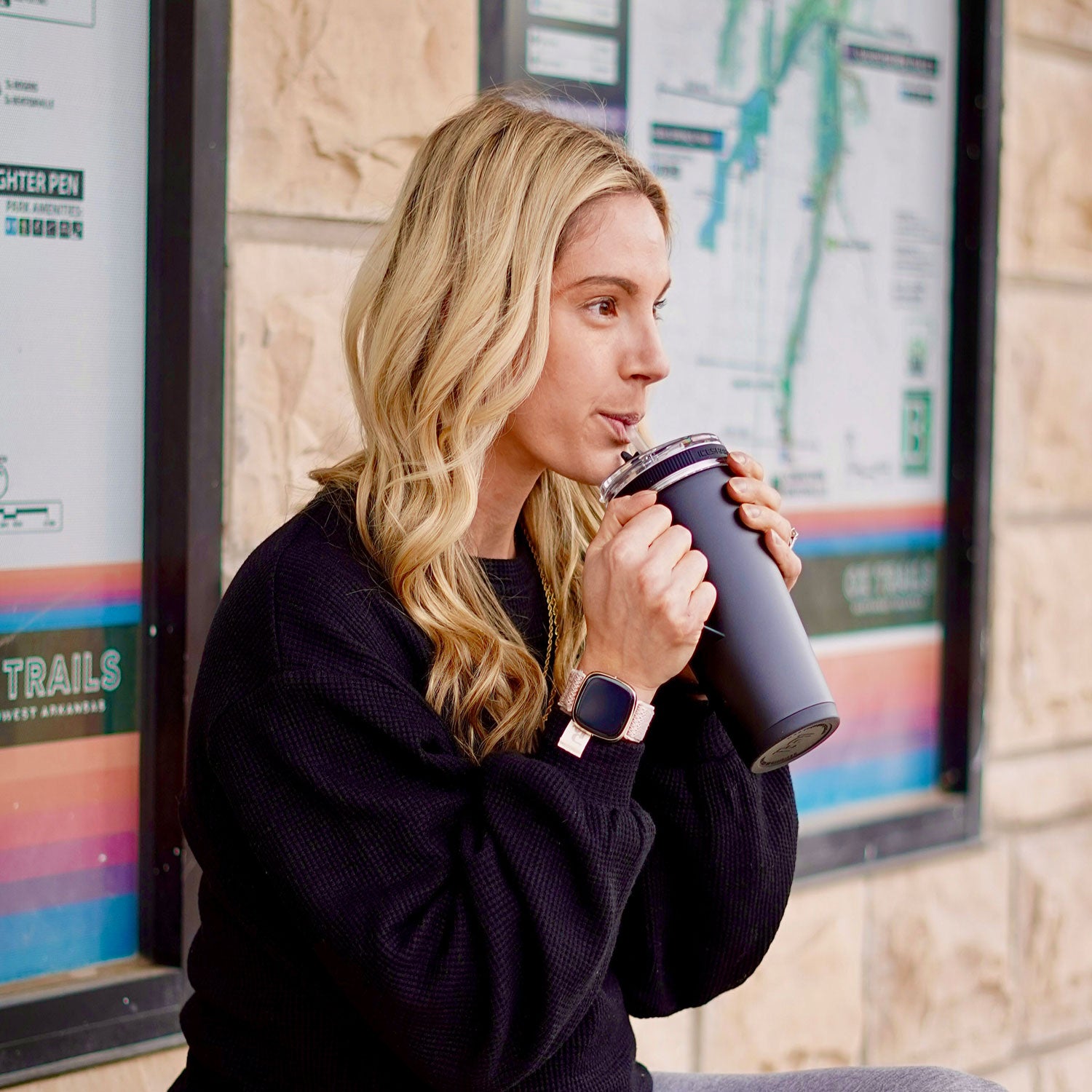 An image of a young, blonde-haired woman sitting outside of a building taking a drink from the black-colored 26oz Flex Bottle with Handle