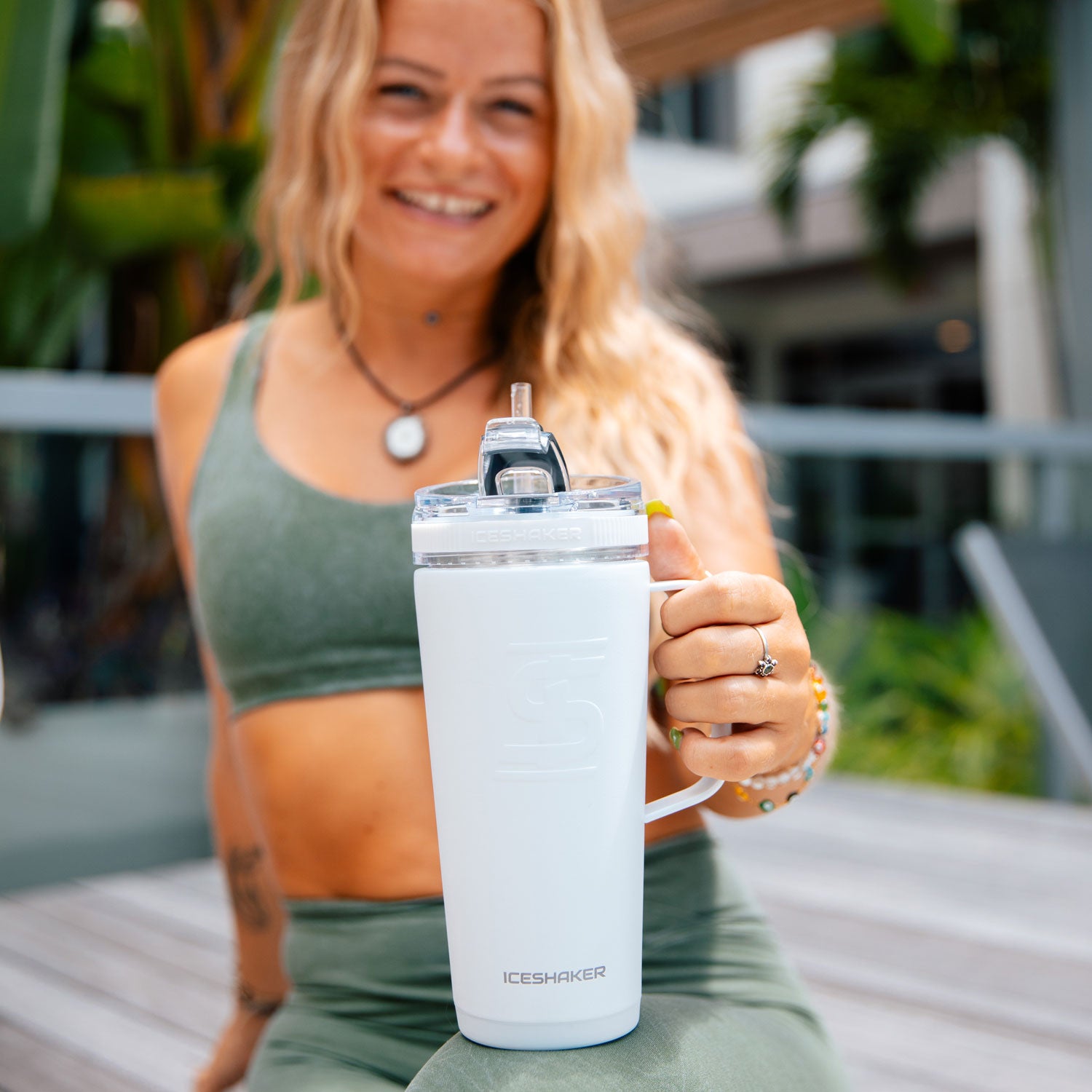 An image of a young woman sitting on a patio holding out a white-colored 26oz flex bottle with a handle.