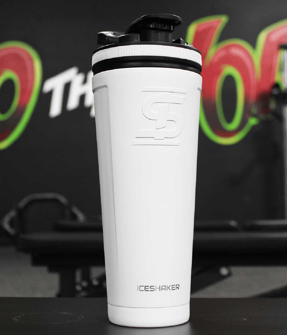 The white-colored 36oz Ice Shaker sits in a gym setting. This image shows the front-view of the bottle.