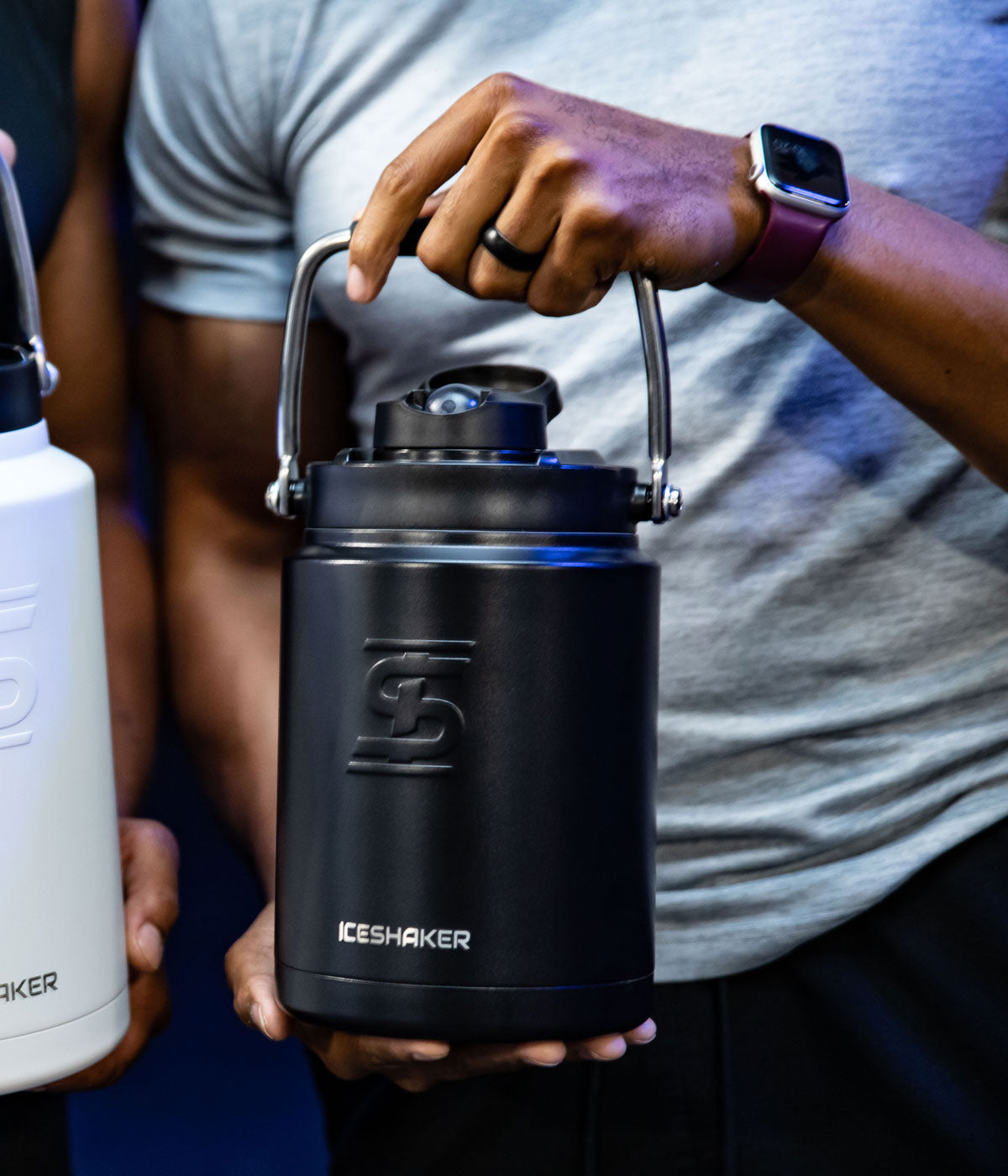 A Black-colored Half Gallon Jug is being held by a set of male hands. One hand is holding the bottom of the jug while the other hand holds the jug by its easy-carry handle.