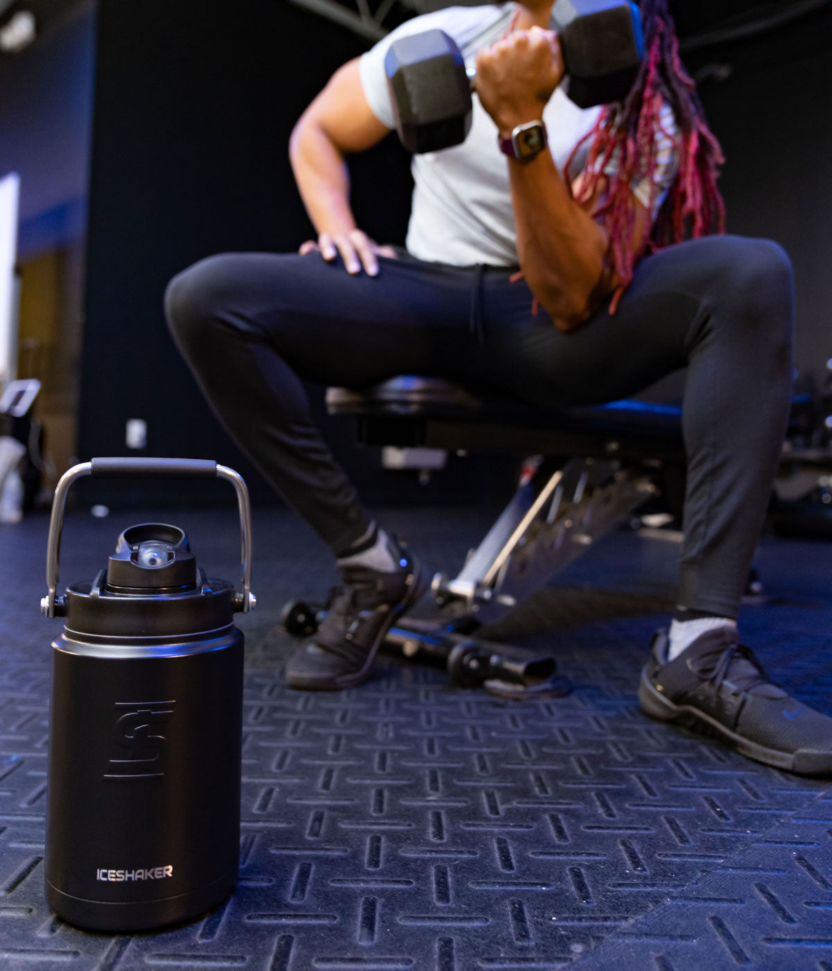 An image of a black-colored Half Gallon Jug sitting on a floor with it's handle up in a gym. In the background, a man is sitting on a workout bench, and lifts a barbell weight with one arm.