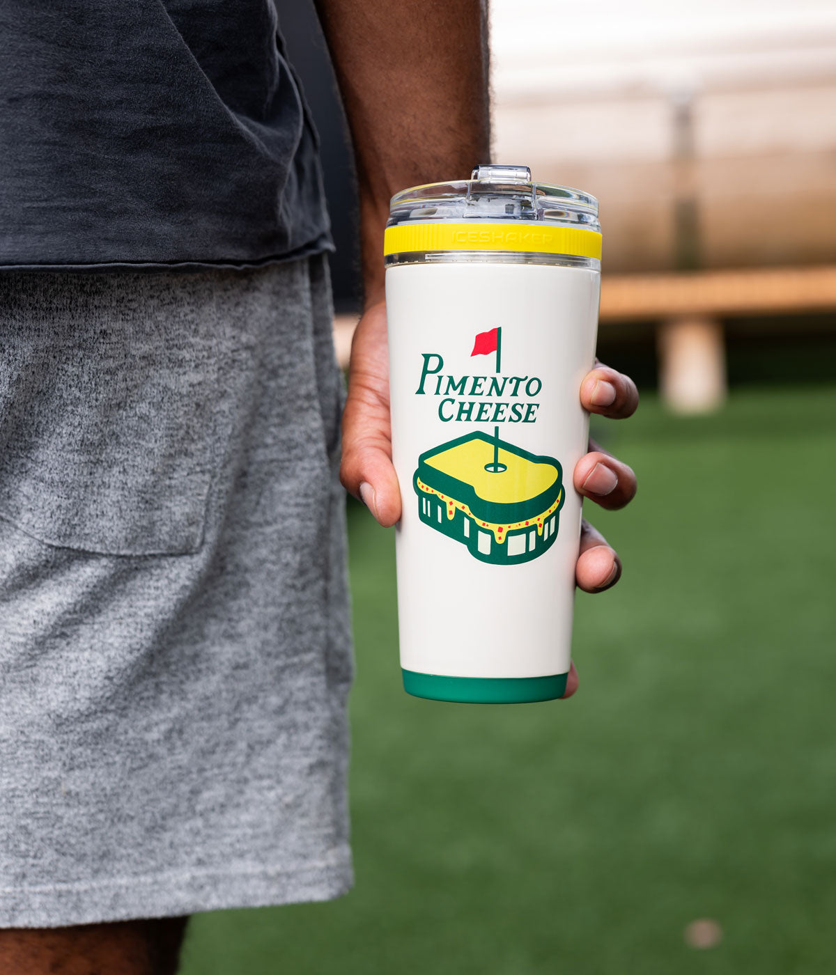 This image shows the Golf Series Pimento Cheese 26oz Flex Bottle being held by a hand. There is greenery in the background.