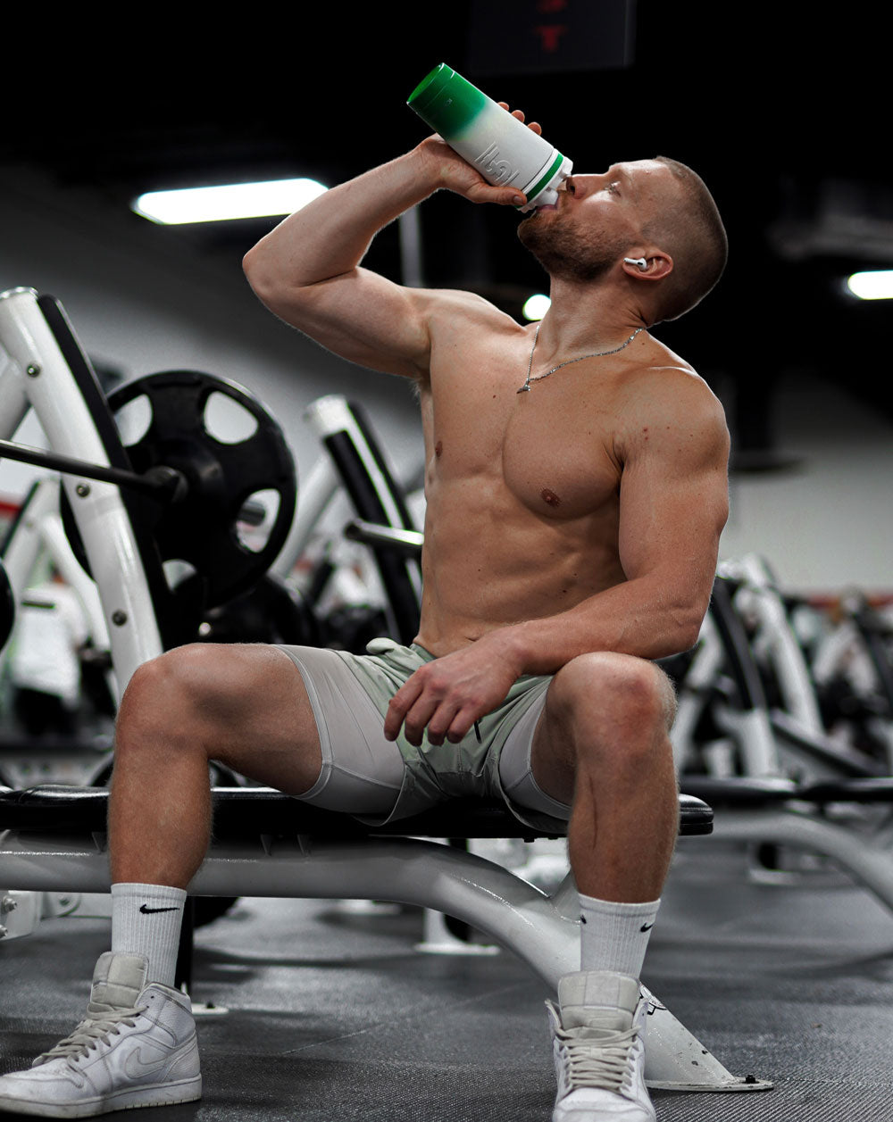 An image of a man with big muscles in a gym. He is sitting on a workout bench and is taking a big drink from his 26oz Ice Shaker.