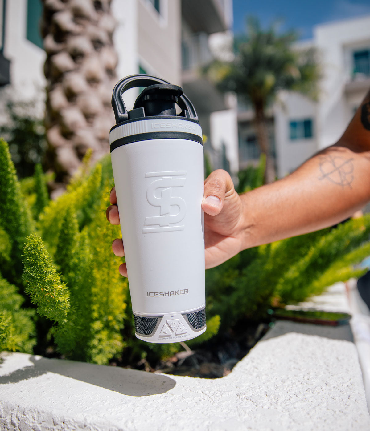 An image of the White 20oz Speaker Bottle being held by a hand. In the background is green plants and a palm tree.