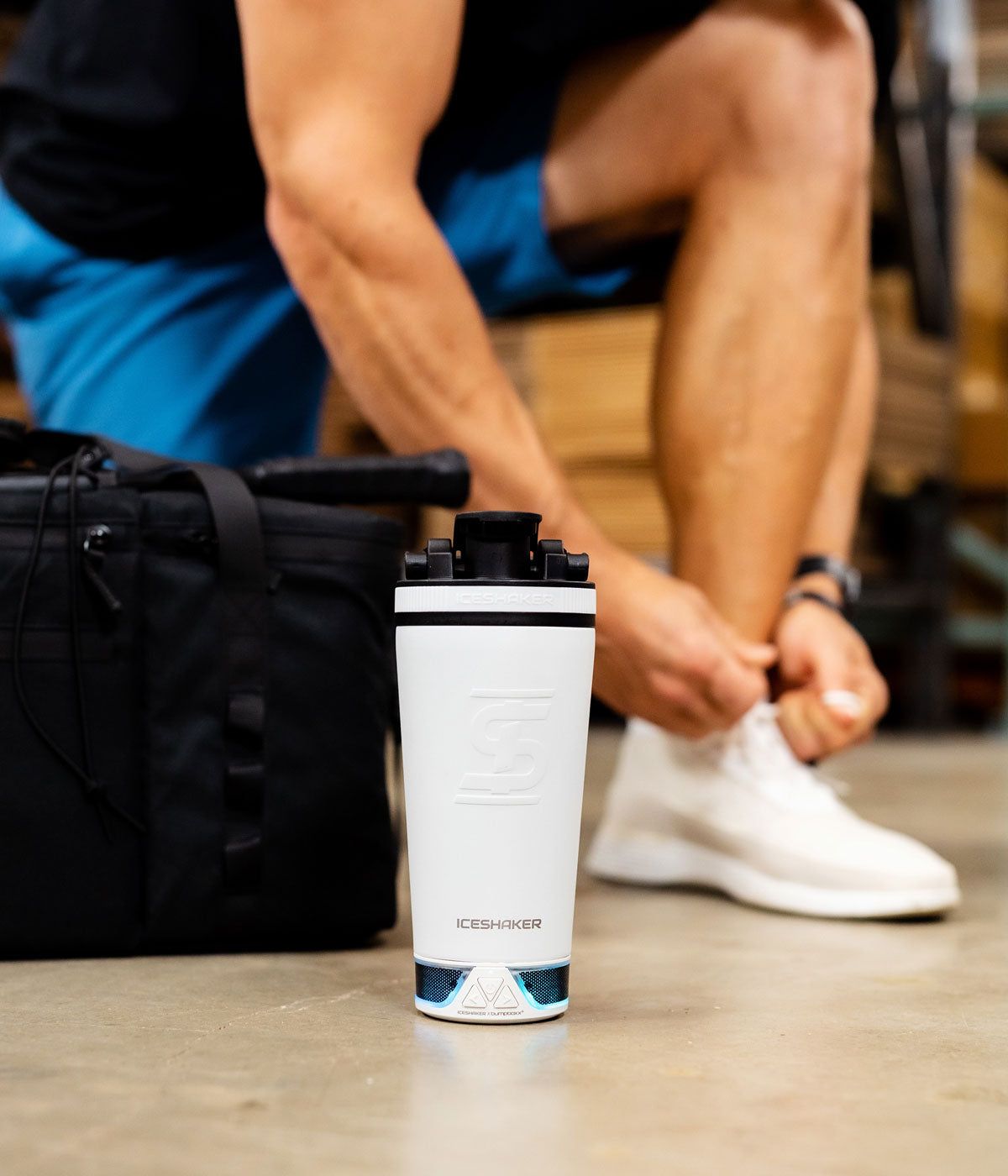 An image of a White 20oz Speaker Bottle with blue lights glowing from the speaker, which indicates that the speaker is on. In the background a man is leaning down tying his shoe.