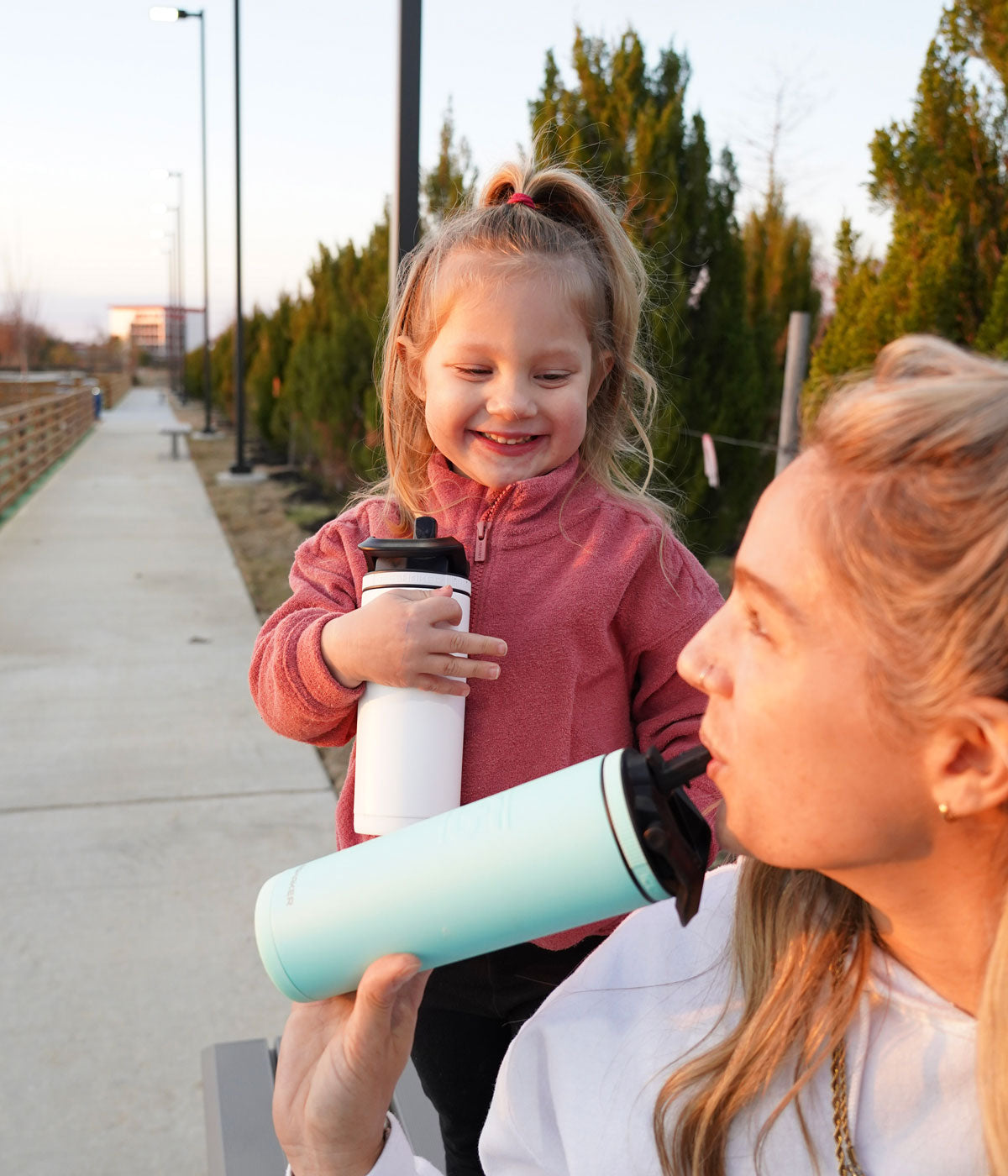 An image of a mom and her daughter with Ice Shaker Sport Bottles. The mom is taking a drink from her Mint 20oz Sport Bottle and looks lovingly at her daughter who is holding a white 14oz Sport Bottle.