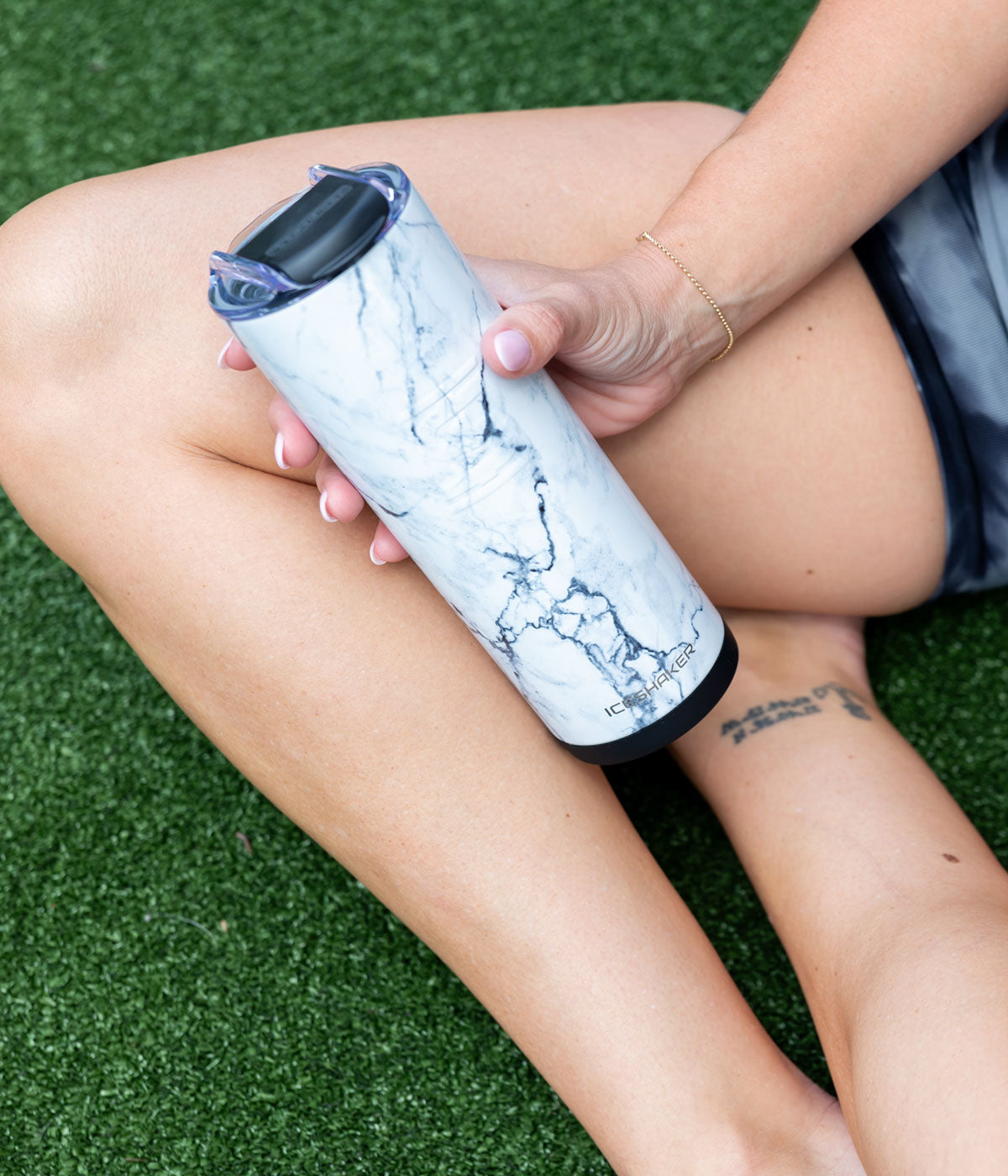 A close up image of the White Marble 20oz Skinny Tumbler. A woman is sitting indian-style on green turn and holds the Tumbler with her hand.