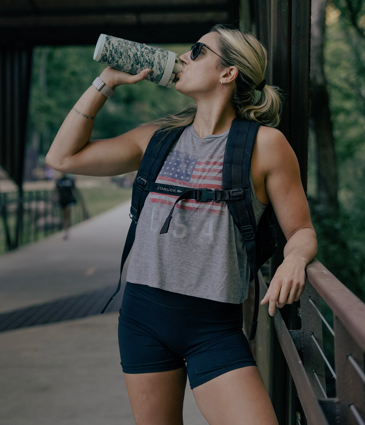 This image shows a young, fit Caucasian woman taking a rest along a concrete walking path. She is taking a refreshing drink from the US Army Camo 26oz Ice Shaker.