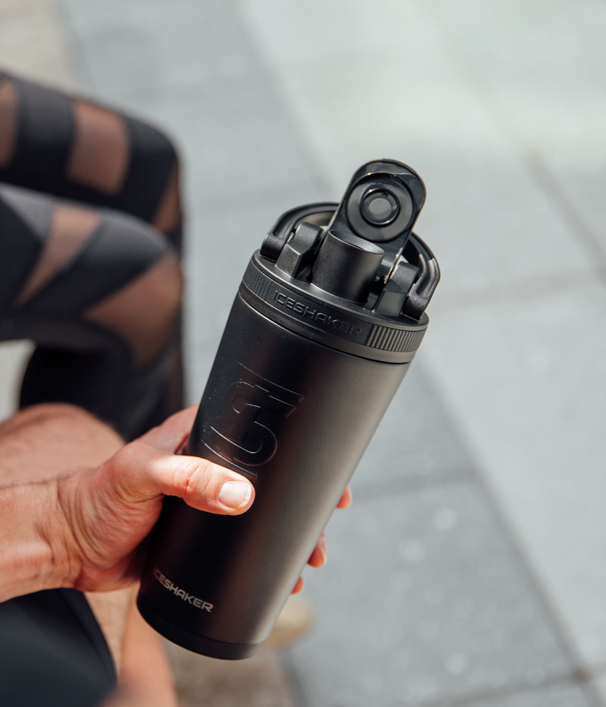 This image shows a close up view of the Black 26oz Ice Shaker. The flip lid top is open and is being held up by a hand.