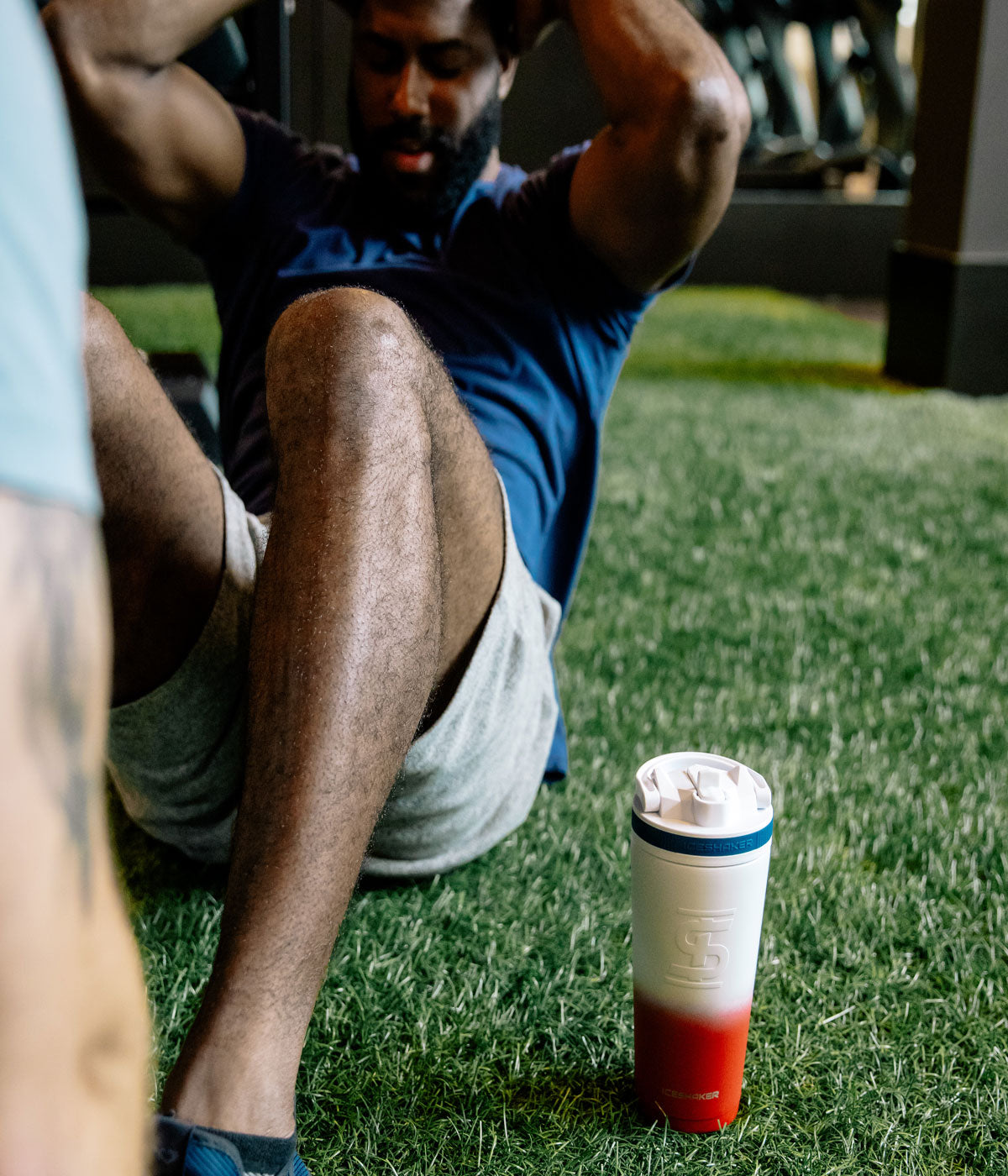 An image of a USA-colored 26oz Sport Bottle sitting next to a man who is doing sit-ups on green turf.