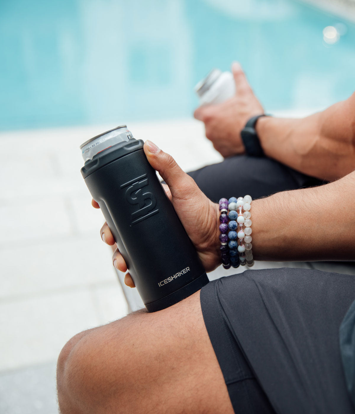 An close up image of the Black 12oz Canpanion being held by a males hand. There is a pool in the background and another man's hand holding a White 12oz Canpanion.
