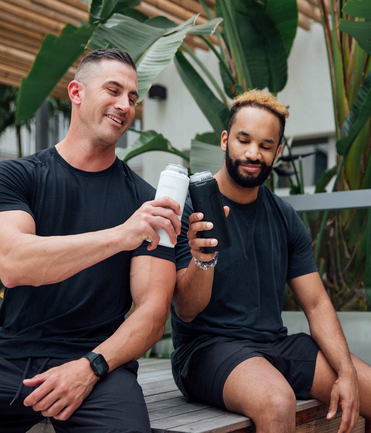 One Caucasian man and one Afican-American man are sitting outdoors. They each have a 12oz Canpanion with a can inserted in their hand and are clinking them together. 