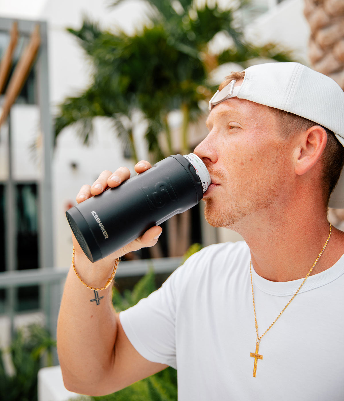 A young Caucasian man wearing a gold Cross necklace and a hat backwards is drinking from a can that is inside a White 12oz Canpanion