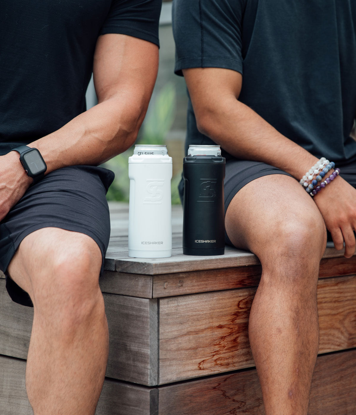 Two men are sitting next to each other on a wooden platform. A white and a black 12oz Canpanion are sitting in between the two men.