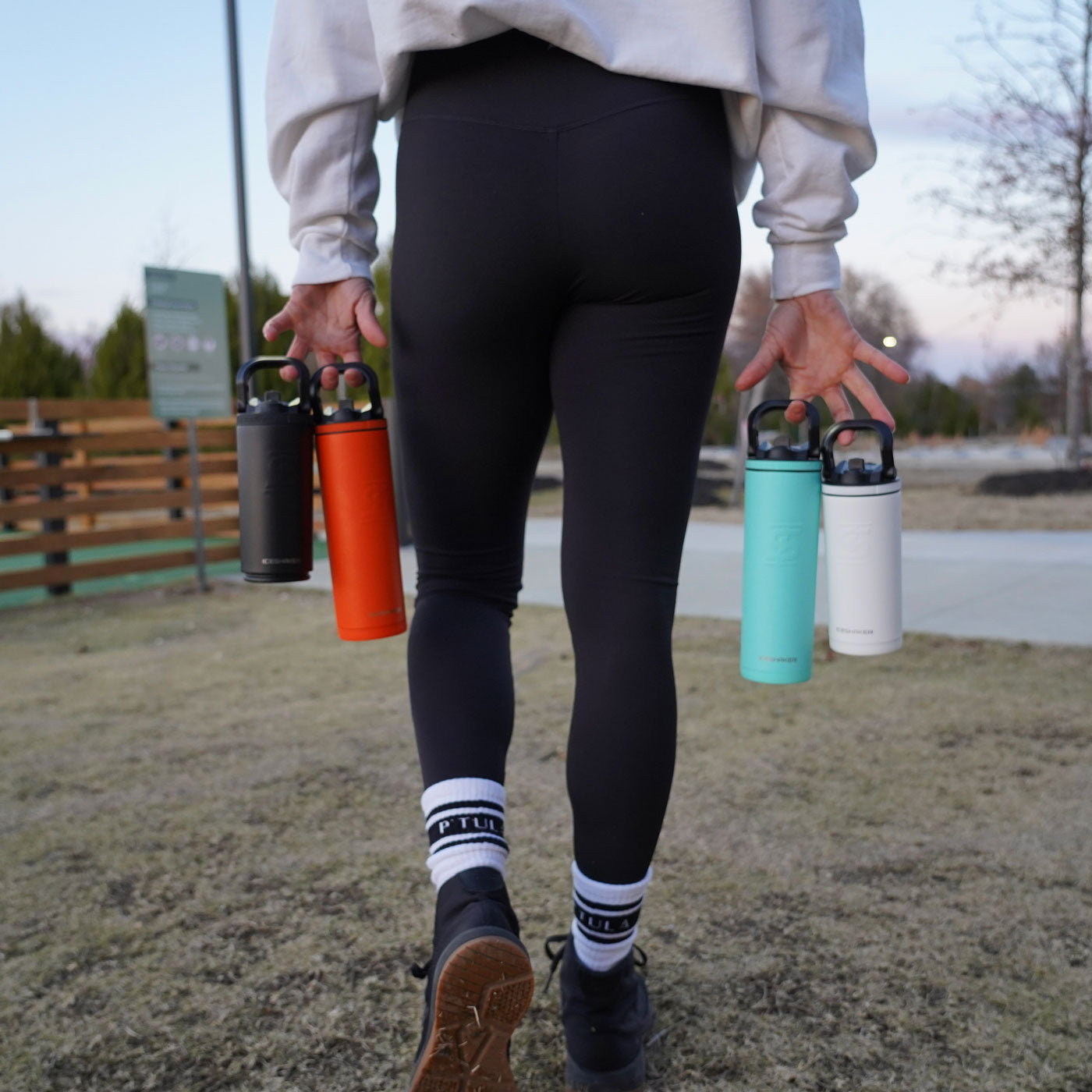 This image shows four different colored Sport Bottles being carried by their handles. A woman is holding them with both of her hands and she is walking away from the camera.
