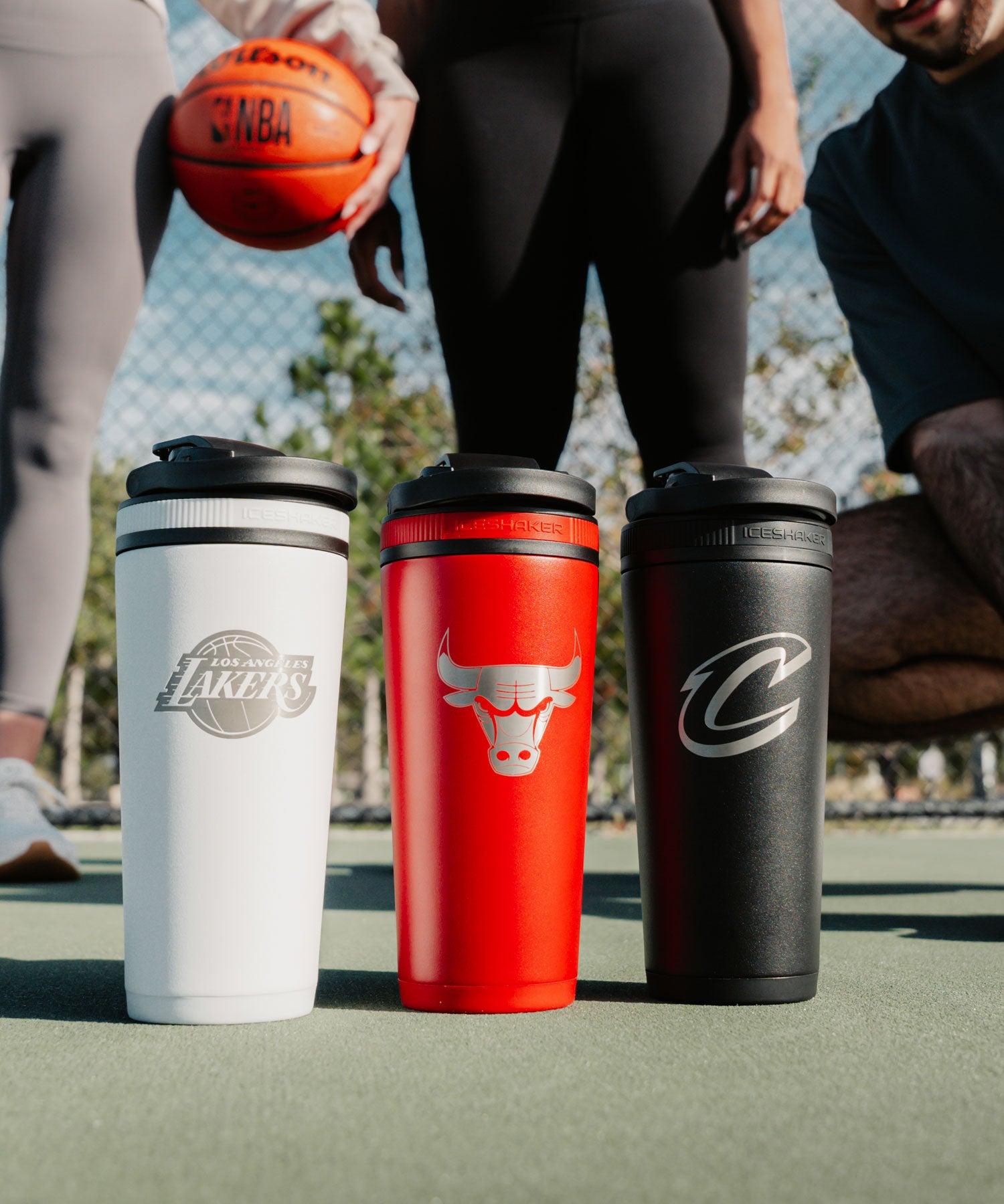 3 engraved NBA 26oz Ice Shakers are lined up next to each other on a basketball court. The sun is shining on the insulated protein shaker bottles highlighting the engraved NBA teams logos on each bottle.