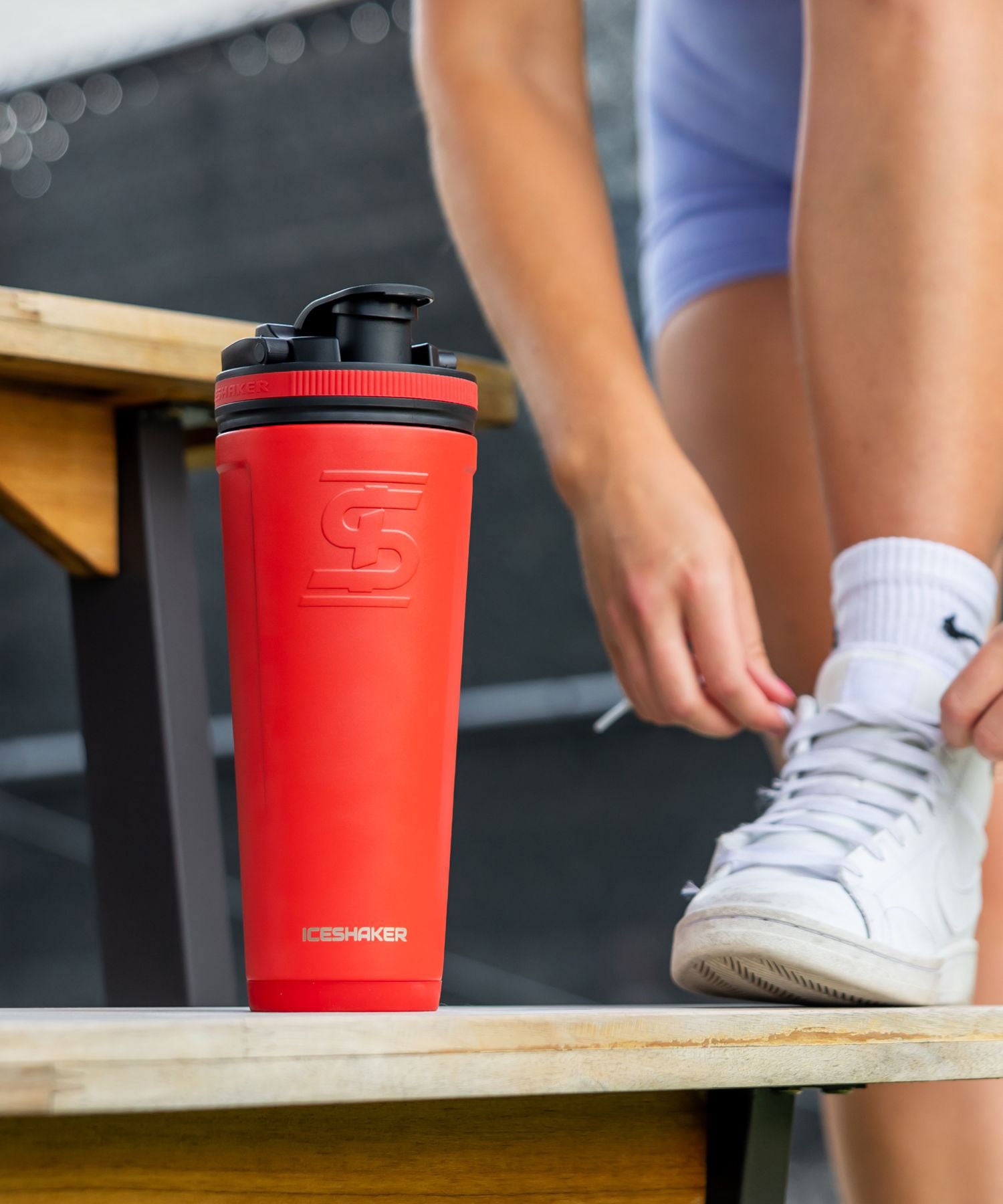 An image of a Red 26oz Ice Shaker sitting on a picnic table bench. A lady has her foot up on the bench next to the Ice Shaker and is tying her white athletic shoe. Click now to Shop 26oz. Ice Shaker Bottles