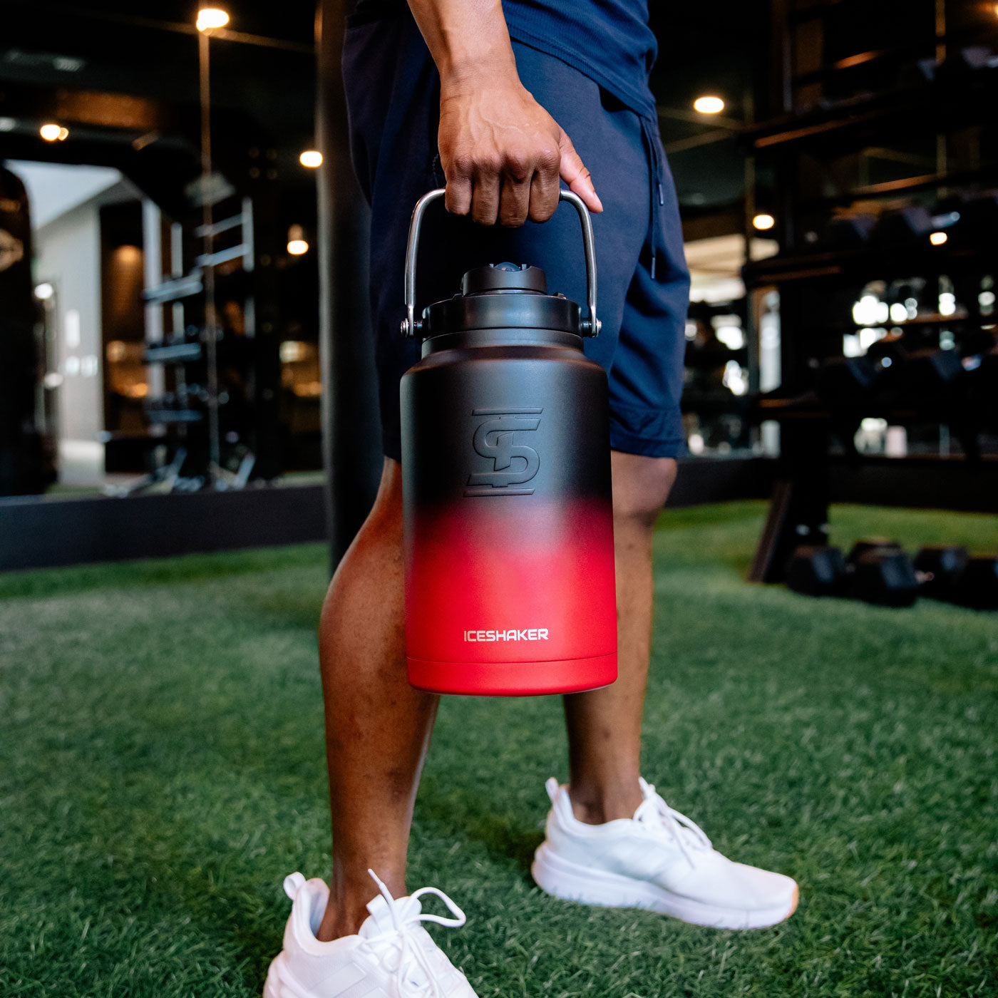 This image shows a red black ombre-colored One Gallon Jug in a gym setting. It's being held by the handle that's built into the lid of the Jug.