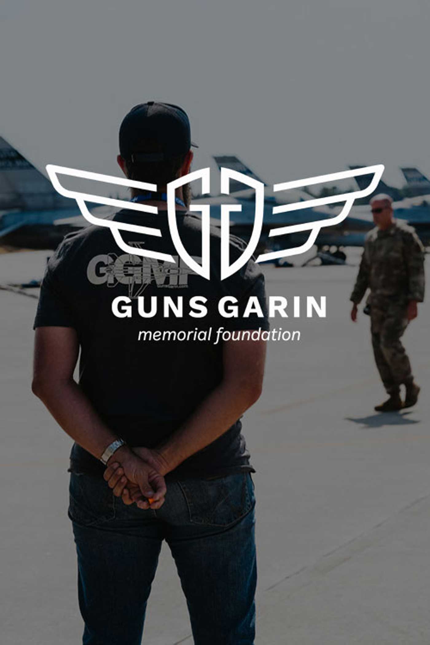 A man wearing a Guns Garin Memorial Foundation t-shirt stands with his hands clasped behind his back as he looks towards jets. The Guns Garin Memorial Foundation is overlayed on top of the image.