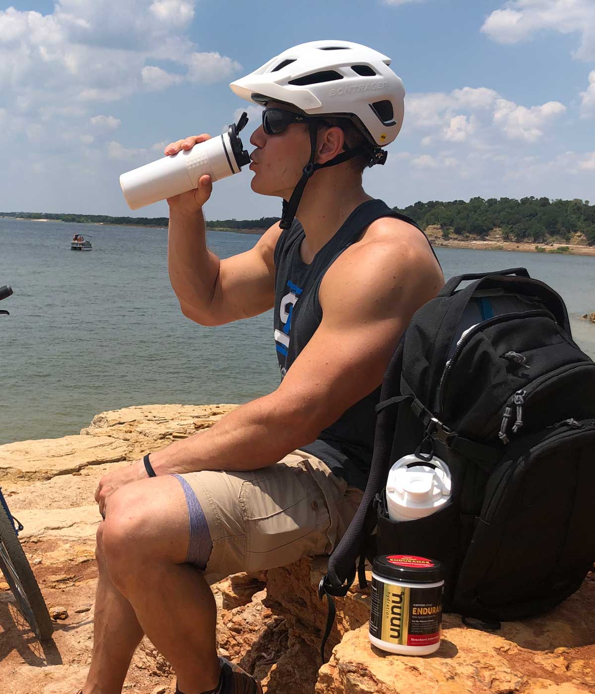 An athletic man wearing a helmet and sunglasses taking a drink from a white 20oz Skinny Shaker. He is sitting on a rock and there is a lake in the background.