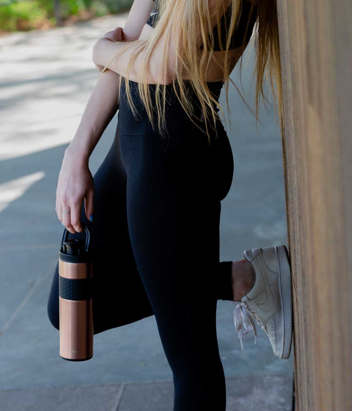 an image of a woman holding the Rose Gold 20oz Skinny Shaker by it's handle that's built into the lid. She is leaning against a wall with one foot propped up and the other on the ground
