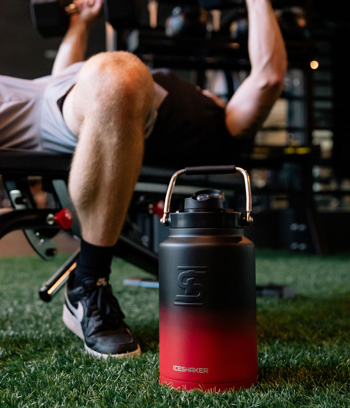 An image of a Red Black Ombre One Gallon Jug sitting on green turf inside a gym. In the background, a man is lifting weights.