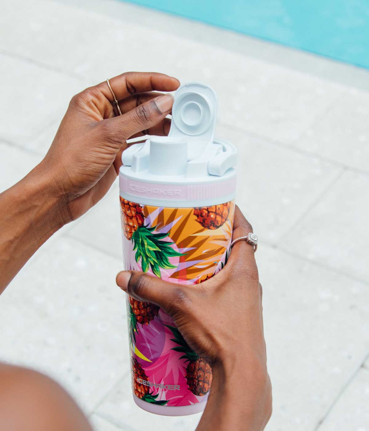 This image shows the Pineapple 26oz Ice Shaker being held by two feminine hands. One hand is lifting up the flap on the flip lid to take a drink from. A pool setting is in the background of the image.