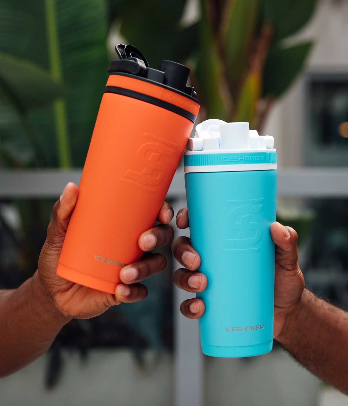 An Orange 26oz Ice Shaker and a Caribbean Blue 26oz Ice Shaker are being held together by two hands. This image shows a close up front view of the 26oz Ice Shakers