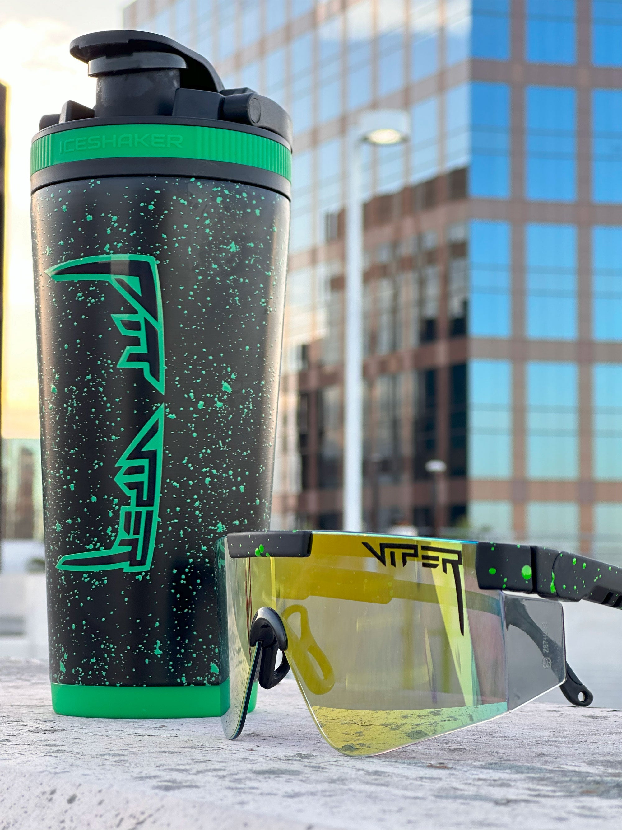 The Custom Pit Viper 26oz Ice Shaker and Pit Viper Sunglasses are next to each other on a concrete platform. Behind them are high-rise buildings.