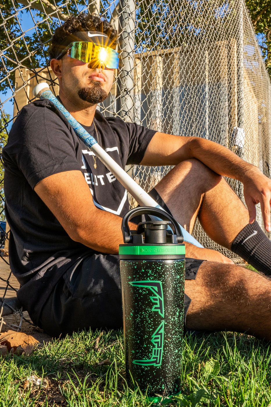 This image shows a young, hispanic man sitting up against a chain-linked fence. The sun is shining bright on him as he wears the Ice Shaker X Pit Viper Sun Glasses. He holds a baseball bat in his hand and next to him is the Custom Pit Viper 26oz Ice Shaker.