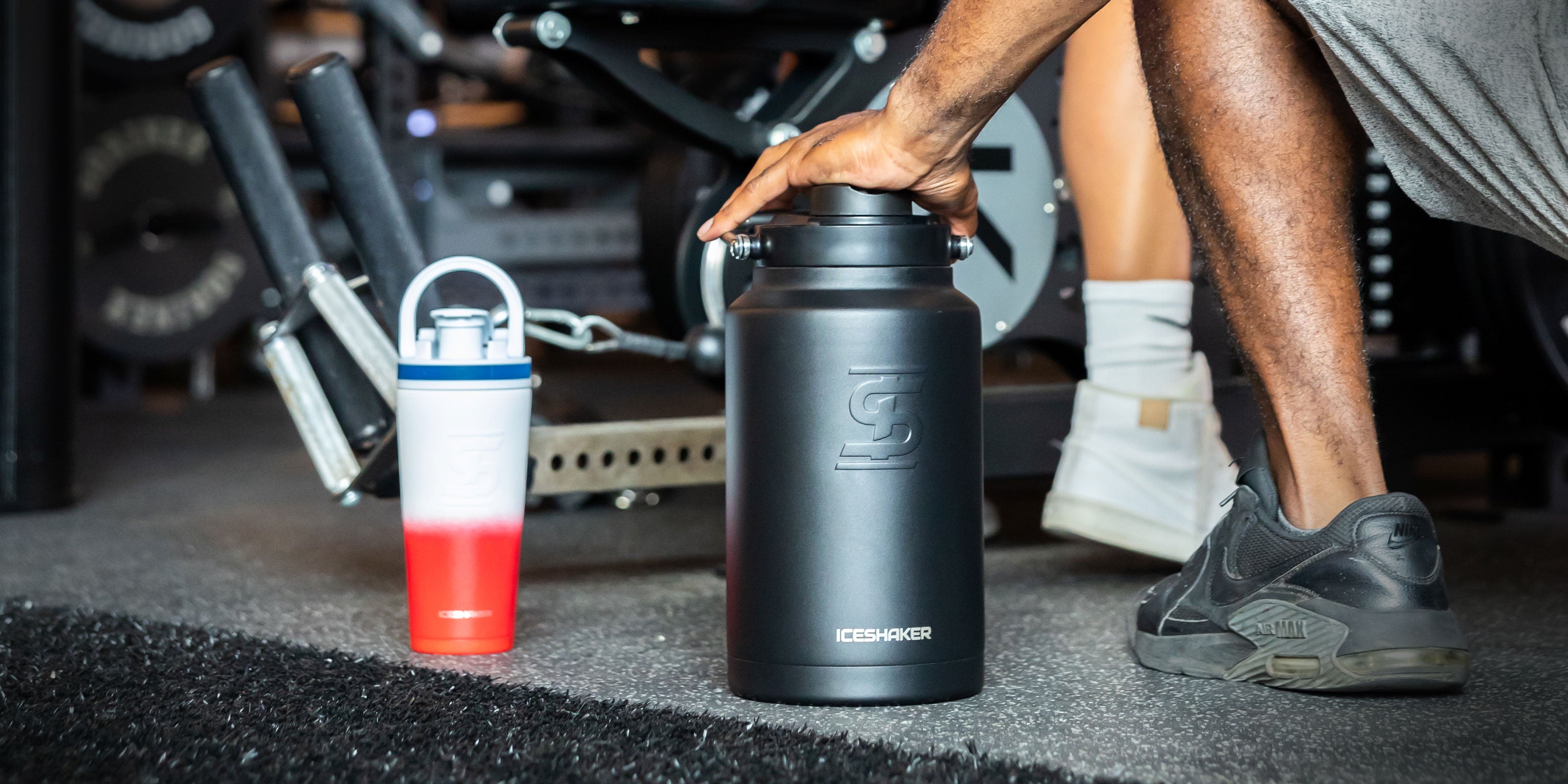 An image of a Black Ice Shaker One Gallon Jug and USA-colored 26oz Ice Shaker Bottle sitting on a gym floor as people workout in the background.