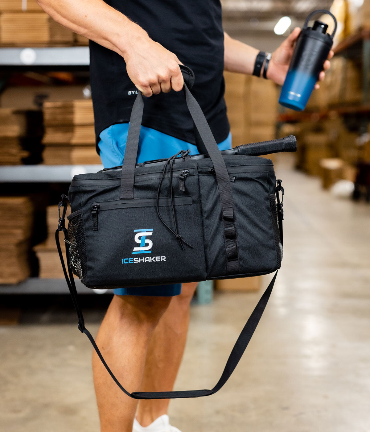 A man holds Ice Shaker's The Duffel Bag by it's durable carry handles in one hand. In the other hand is a Navy Black Ombre-colored 26oz Ice Shaker