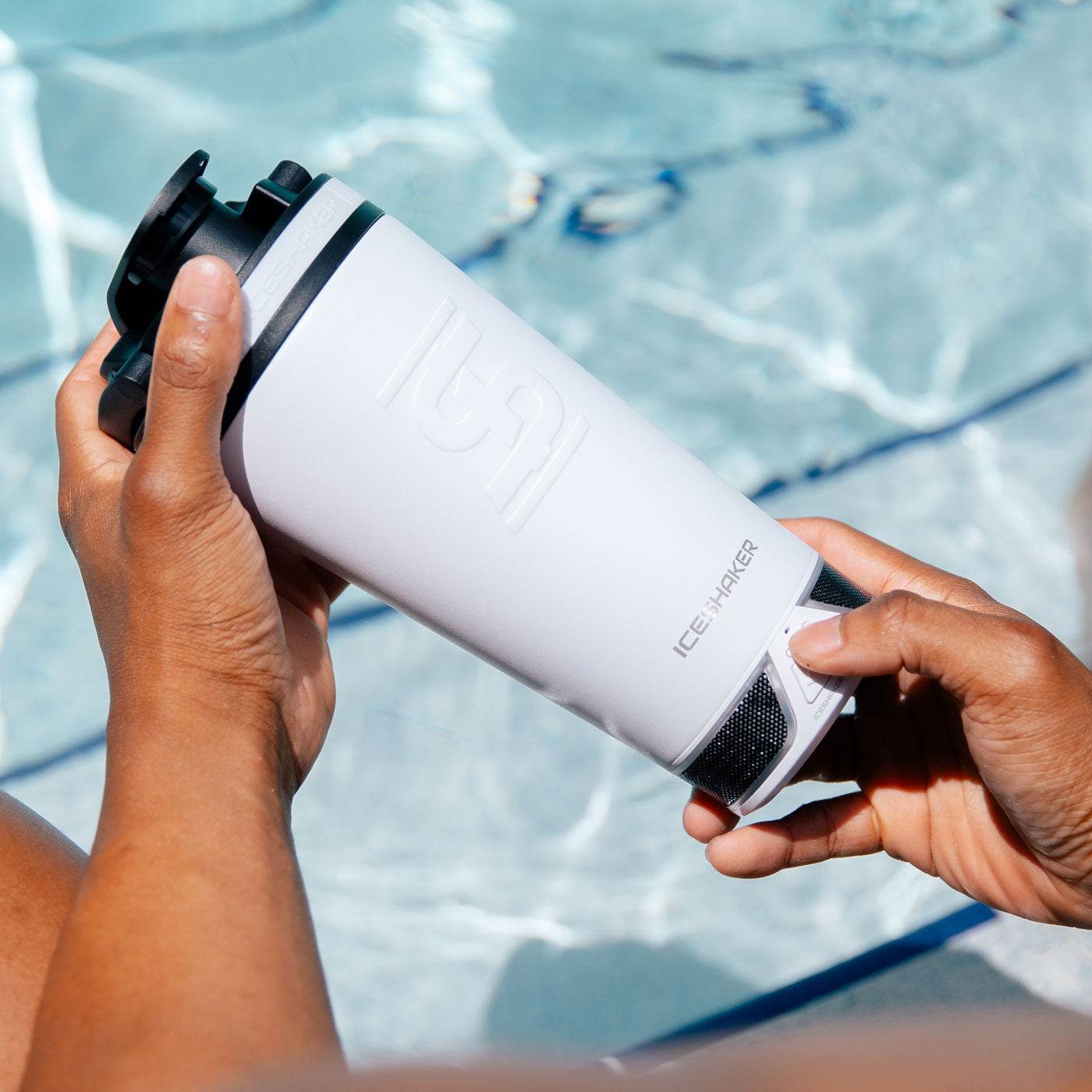 an image of the White 20oz Speaker Bottle being held by two hands. One hand is pressing the power button on the Bumpboxx Speaker that's twists on the bottom of the 20oz Speaker Bottle. There is a pool in the background of the image.